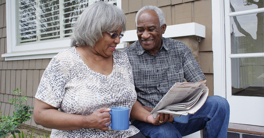 An older African American couple sits outside of their home reading the newspaper together
