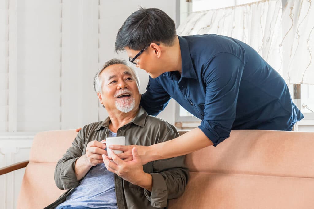 An older Asian man receives a coffee cup from his son as they sit on the couch of their home