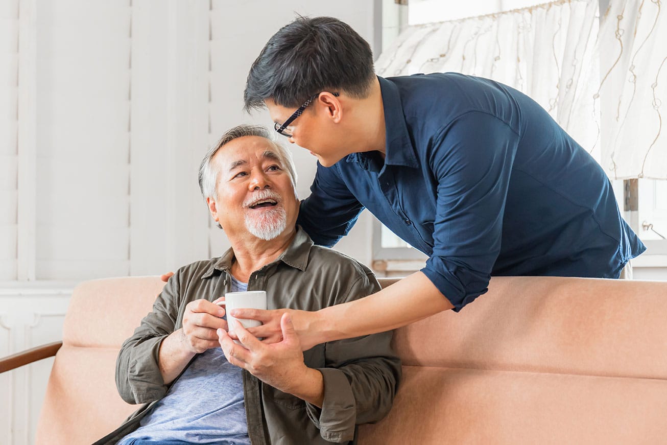 An older Asian man receives a coffee cup from his son as they sit on the couch of their home