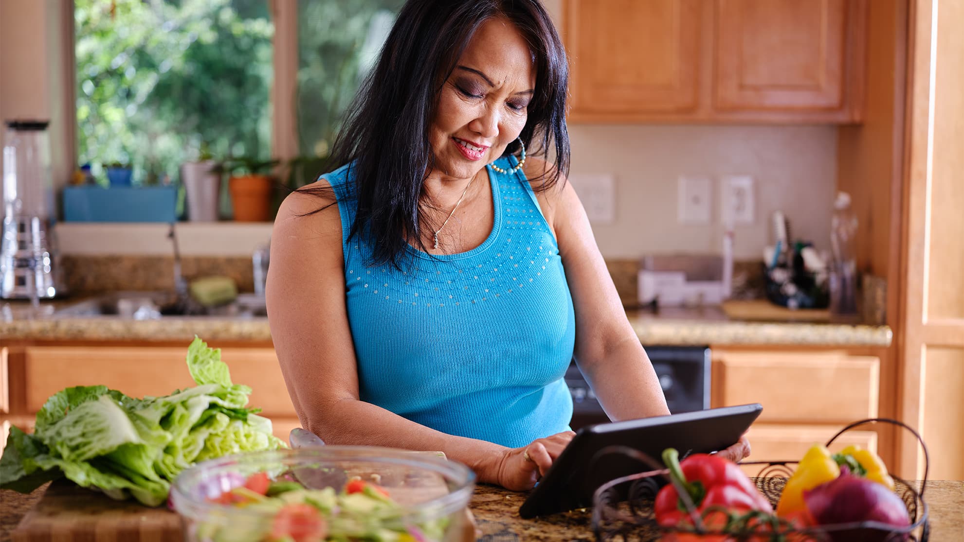 older adult with hispanic or mexican heritage in her kitchen looking at an ipan. On the counter, healthy vegetables can be seen, as well as, a basket with peppers inside.