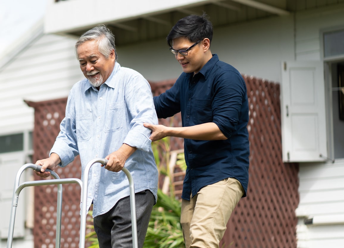 An older adult with his son helping him outside as he uses a walker