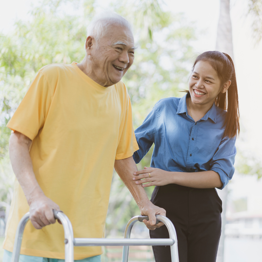 A young direct care workers aids an older adult to walk with aid of his walker. Both are smiling.