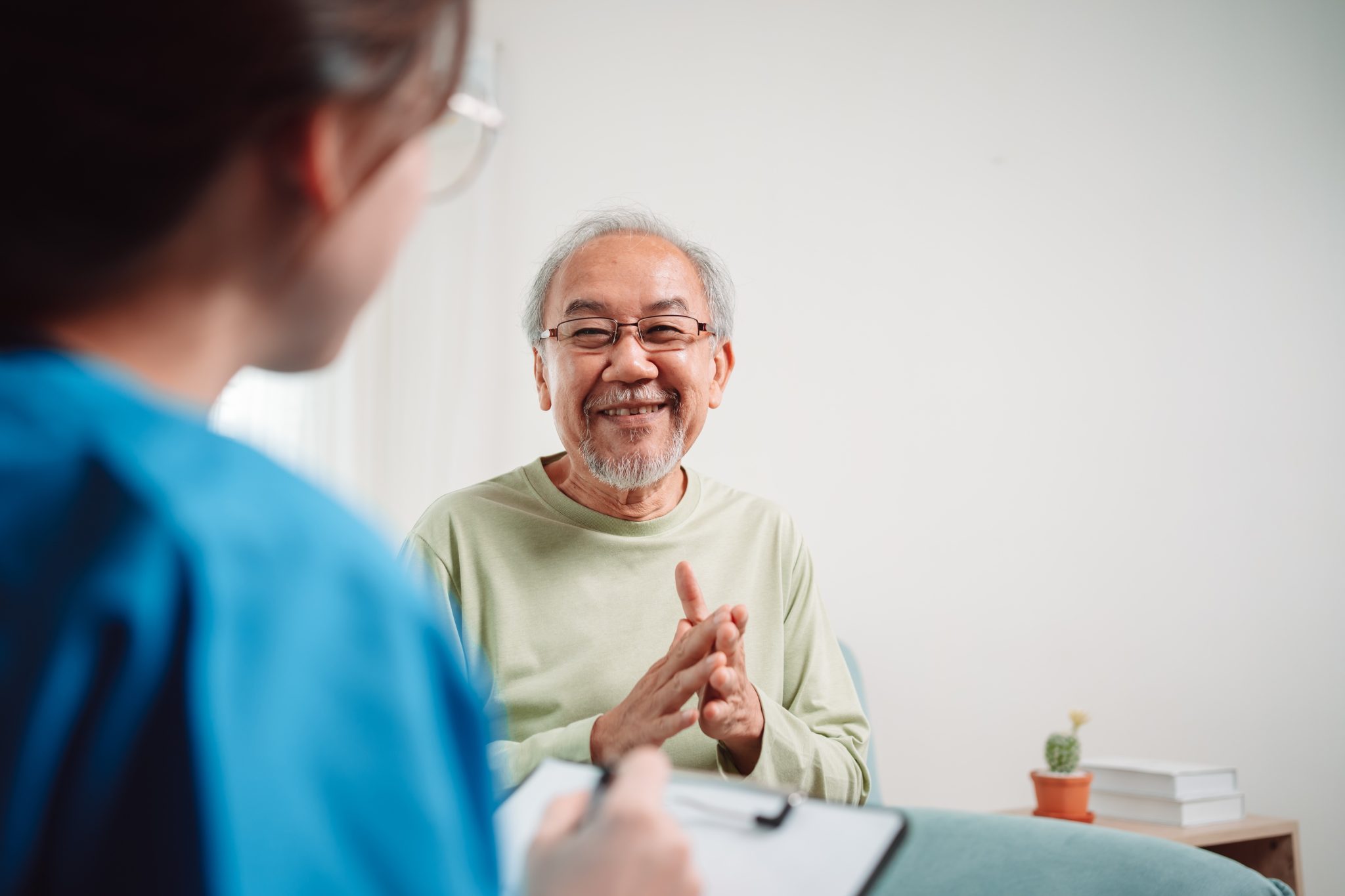 Smiling older man sitting in a chair and speaking with a healthcare professional holding a clipboard during an in-home consultation.