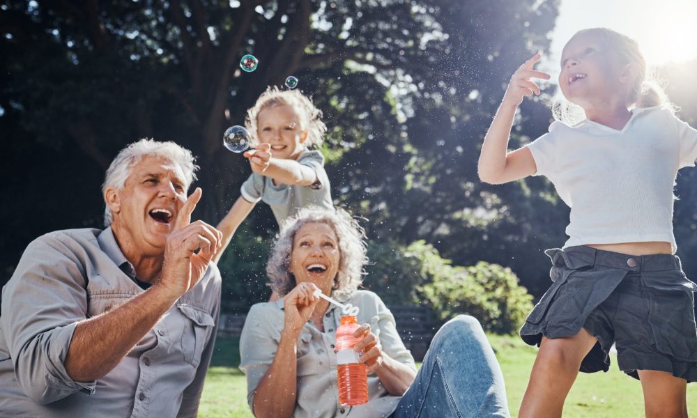 Grandparents, bubbles and children play in park happy together for fun, joy and outdoor happiness. Retired, smile and excited elderly senior couple, girl grandkids and love playing outside in nature.