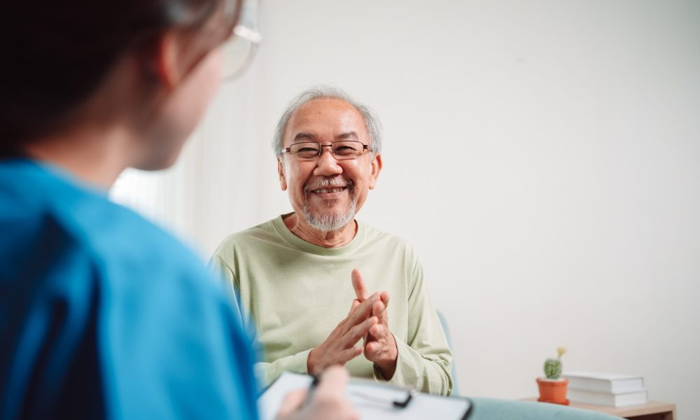 Smiling older man sitting in a chair and speaking with a healthcare professional holding a clipboard during an in-home consultation.